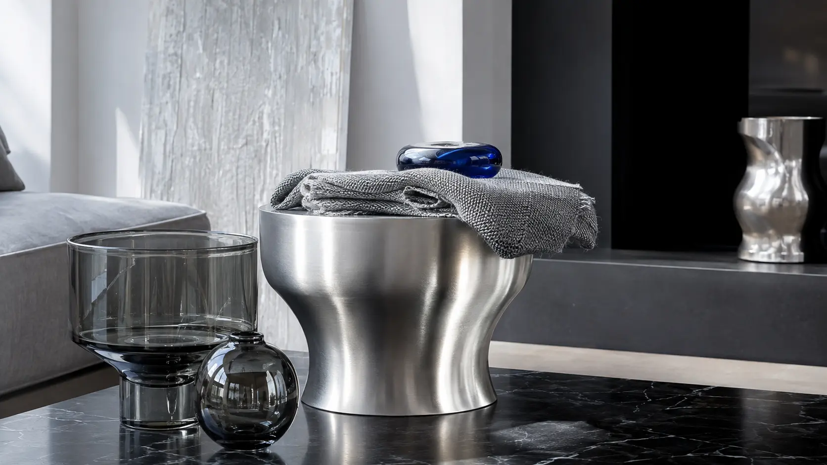 Intérieur contemporain avec table en métal argent brossé, textile gris, verre fumé et objet bleu cobalt
