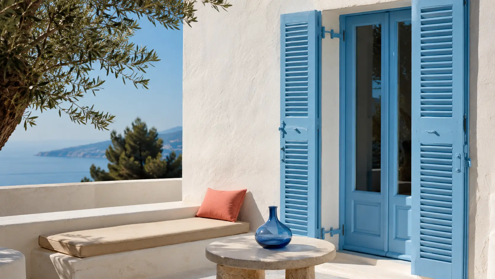 Terrasse méditerranéenne avec volets azur, façade blanche, pierre sable, coussin corail et vase bleu encre