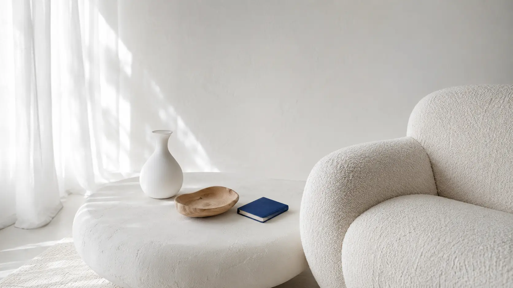 Intérieur blanc avec fauteuil bouclé, table en plâtre, porcelaine, rideau en lin, bois blond et petit livre bleu