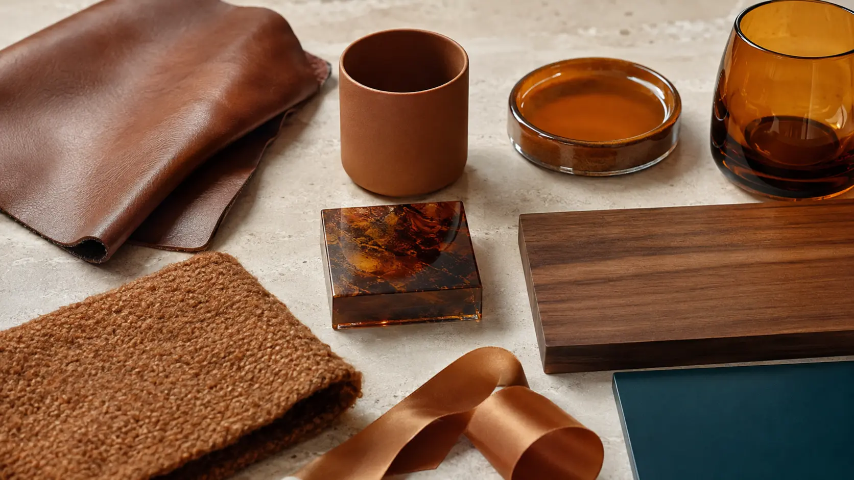 Étude de matières caramel réunissant cuir brun orangé, céramique mate, verre ambré, résine translucide, noyer, laine bouclée, ruban satiné et panneau bleu pétrole sur pierre claire