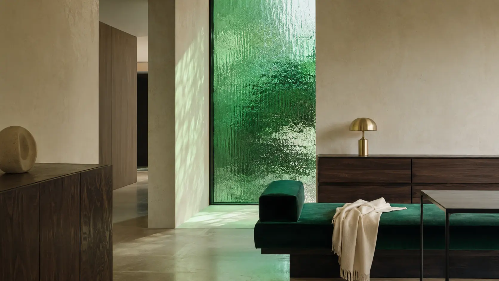 Intérieur contemporain avec panneau en verre texturé vert émeraude, banquette en velours vert, murs de pierre claire, noyer sombre, table en métal noirci et petite lampe en laiton brossé
