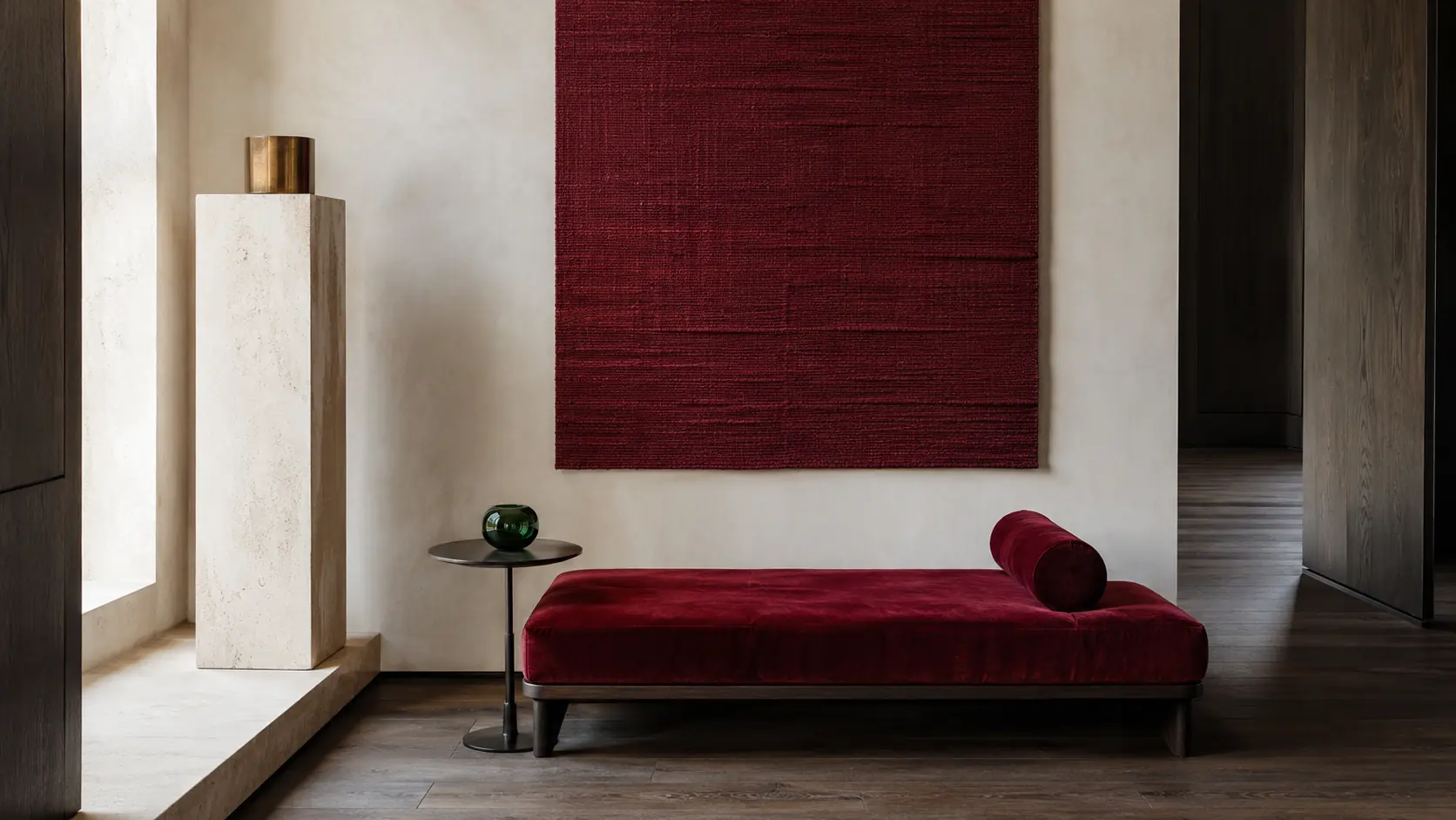Intérieur contemporain avec grand panneau textile grenat, banquette en velours rouge sombre, murs en pierre ivoire, chêne fumé, table en métal noirci et petit vase en verre vert sombre