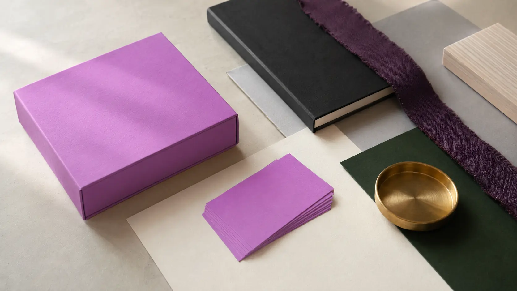 Système d’identité sans marque avec boîte et cartes mauve, papiers ivoire et gris vierges, carnet noir encre, papier vert forêt, bande textile prune fumée, frêne clair et coupelle en laiton