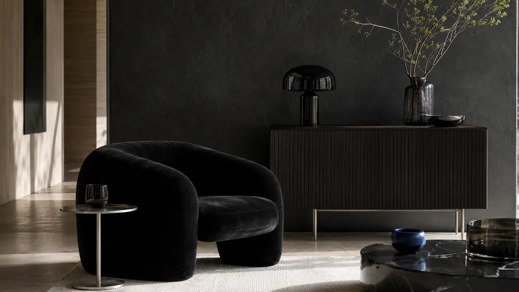 Intérieur contemporain avec grand mur noir en enduit minéral, fauteuil en velours noir, meuble cannelé en chêne noirci, objets en laque noire et verre fumé, acier brossé
