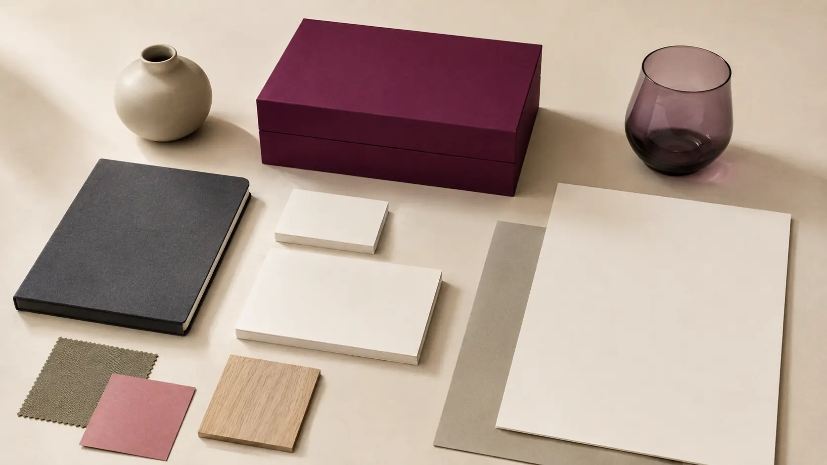 Système d’identité sans marque avec boîte et cartes prune vierges, papiers ivoire, carnet anthracite, feuilles rose poudré et olive, chêne clair, verre fumé et céramique mate