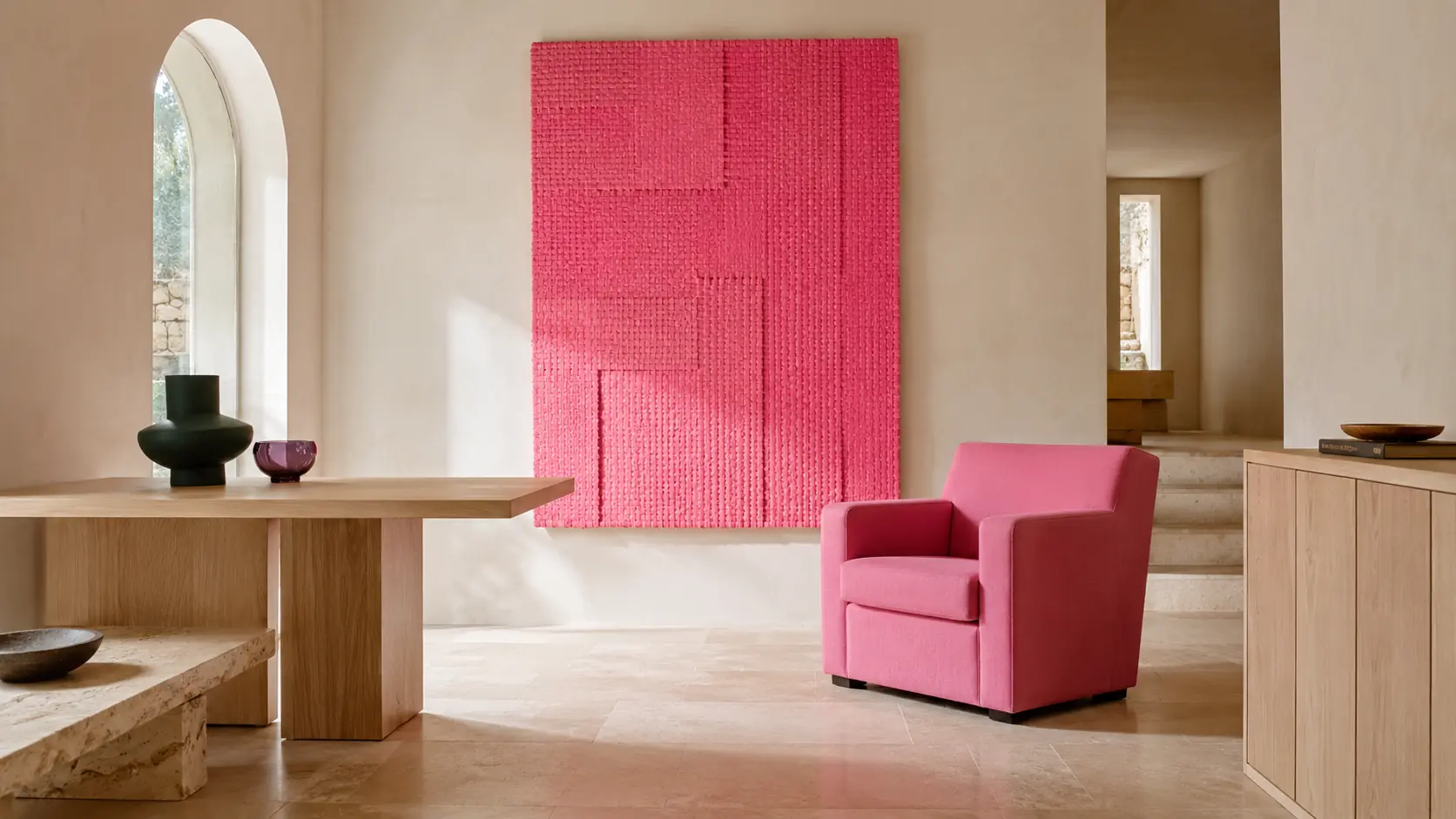 Intérieur contemporain lumineux avec panneau textile rose, fauteuil rose structuré, murs ivoire, mobilier en frêne clair, sol en travertin, céramique vert forêt et petit objet prune