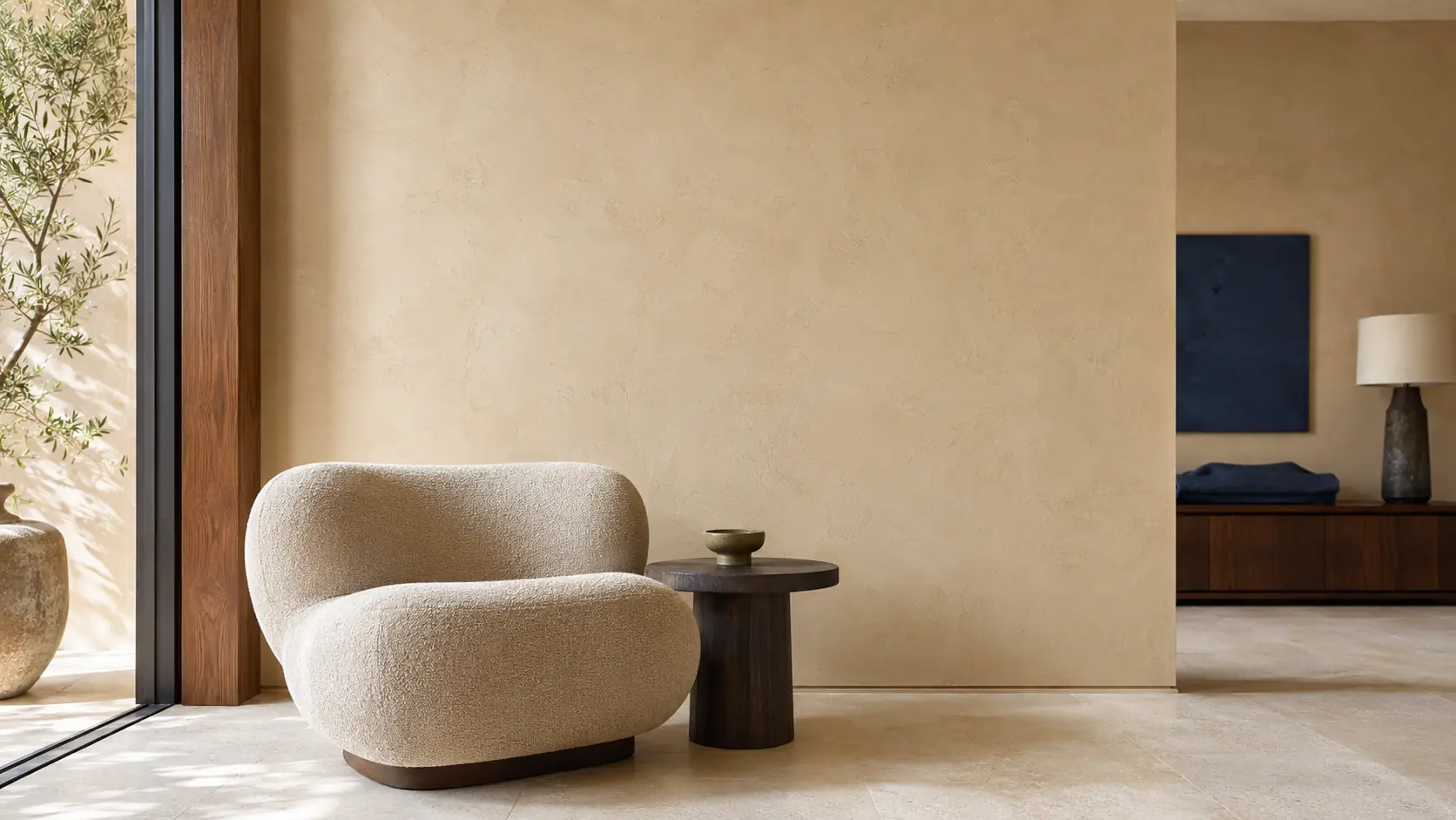 Intérieur contemporain avec grand mur en enduit couleur sable, fauteuil en laine sable, sol calcaire clair, console en noyer, textile indigo et petite céramique olive