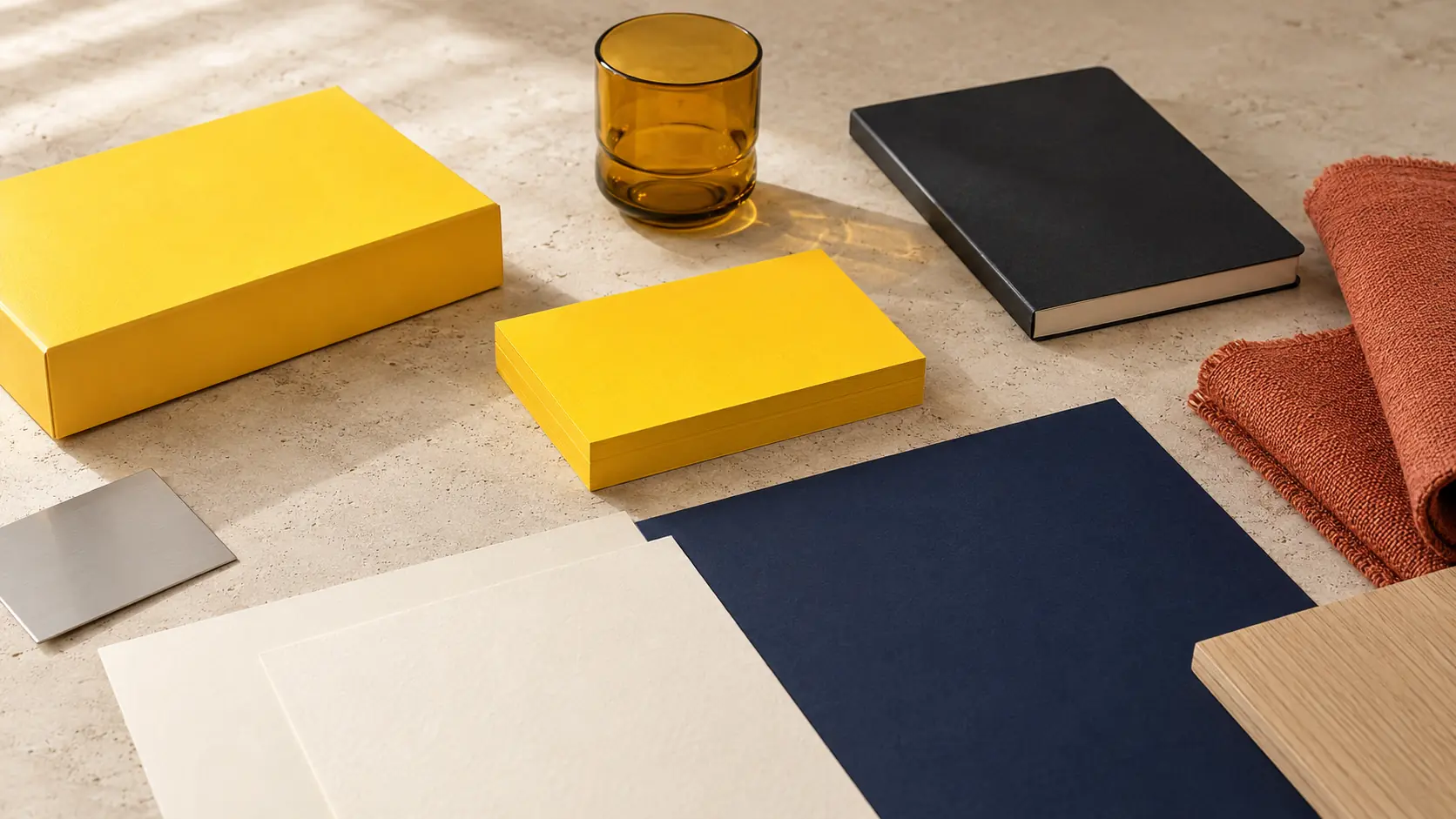 Système d’identité sans marque avec boîte et cartes safran vierges, papiers ivoire, carnet encre, grande feuille bleu nuit, textile terre cuite, chêne clair, verre ambré et métal brossé