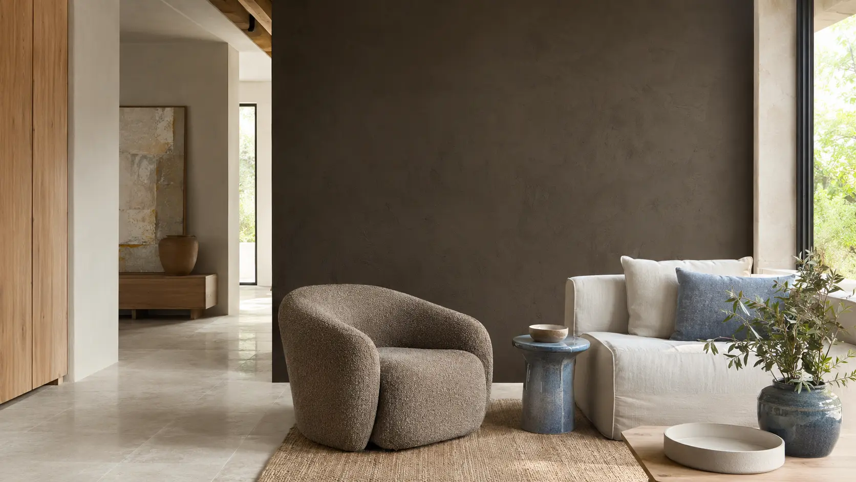 Intérieur contemporain lumineux avec grand mur en enduit taupe sombre, assise en laine taupe, sol calcaire, chêne clair, textile écru et objet bleu grisé