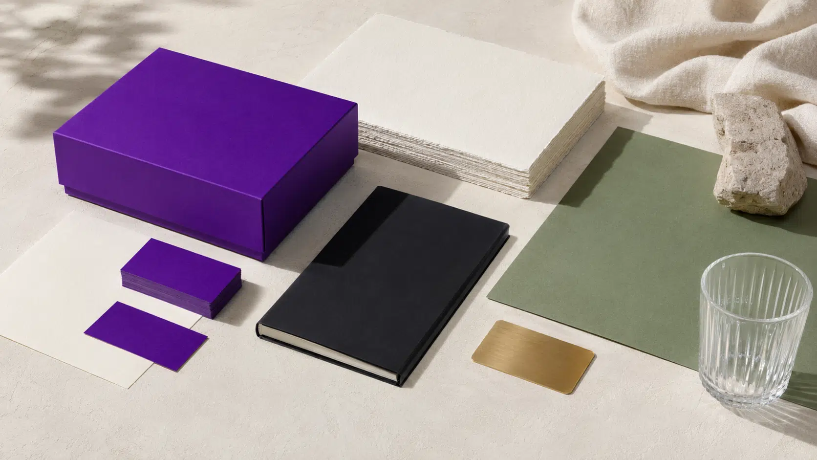 Système d’identité sans marque avec boîte et cartes violet sombre vierges, papiers craie, carnet encre, grande feuille vert lichen, laiton mat, verre clair et textile ivoire