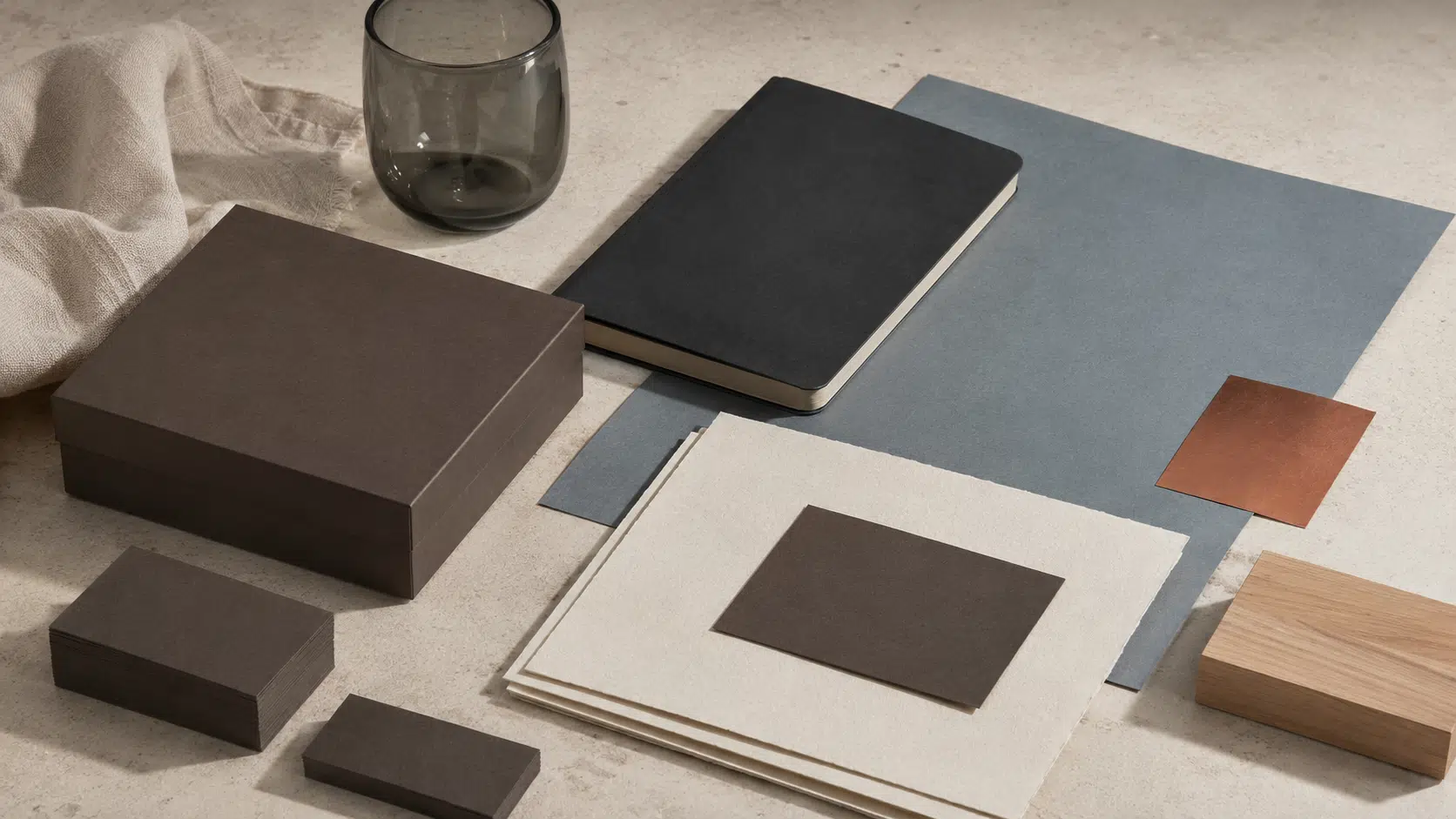 Système d’identité sans marque avec boîte et cartes wengé vierges, papiers calcaire, carnet encre, grande feuille bleu grisé, cuivre mat, lin écru, verre fumé et bois clair